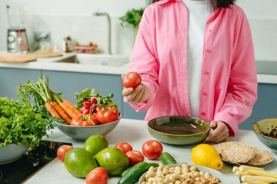 Woman in pink overshirt holds tomato in kitchen with fresh fruits and veggies.